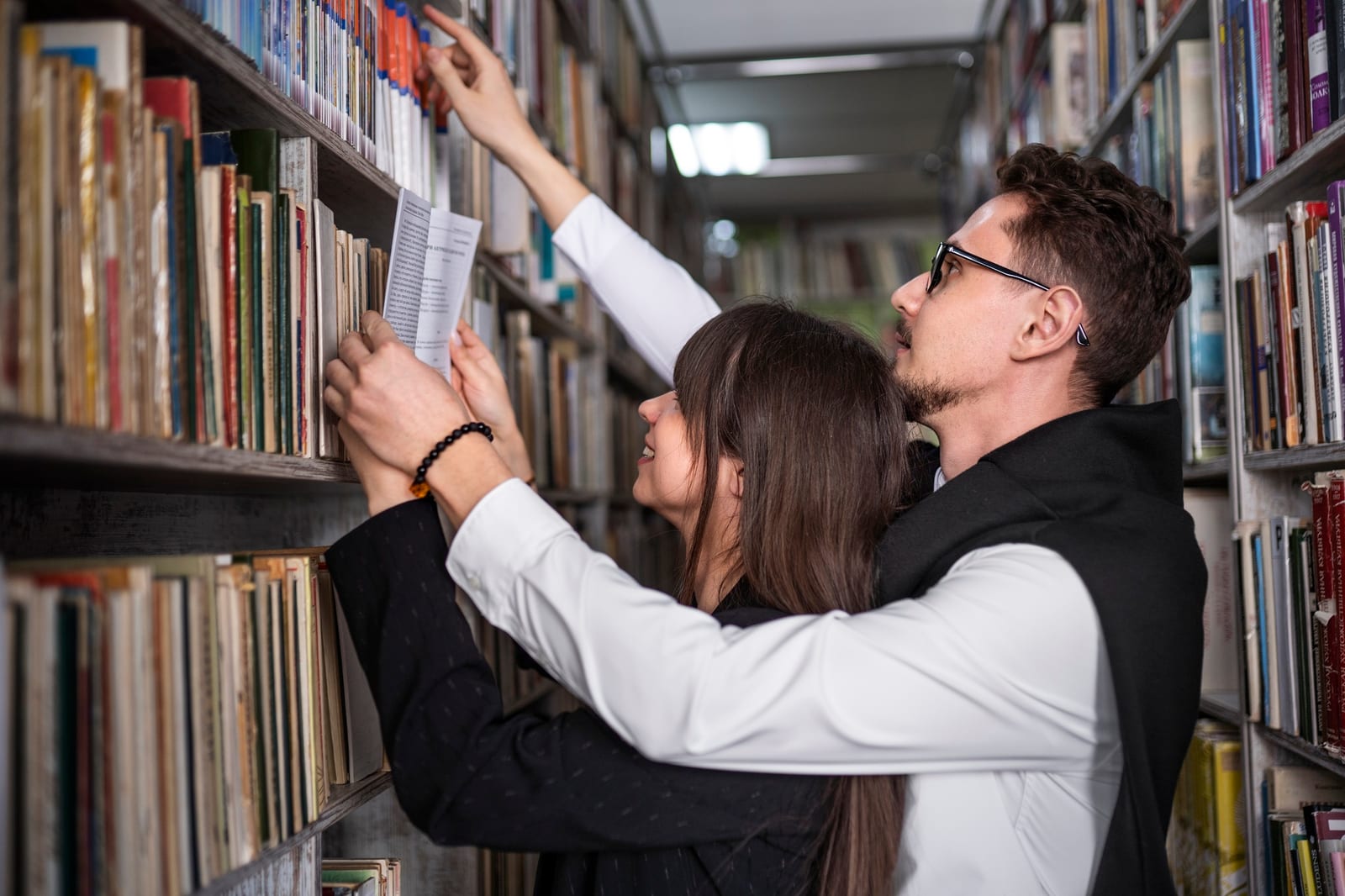 Duas pessoas em um corredor de estantes, buscando e apontando para livros nas prateleiras.